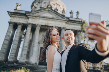 Gorgeous bride and groom taking selfie on background of old castle in park. Stylish wedding couple hugging, smiling and holding phone for selfie photo