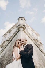 Gorgeous wedding couple embracing in sunlight near old castle in beautiful park. Stylish beautiful bride and groom gently hugging on background of ancient building and nature