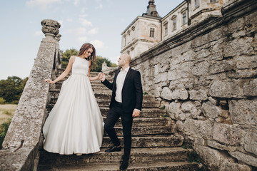 Gorgeous wedding couple walking on stone stairs near old castle in park. Stylish beautiful bride in amazing gown and  groom posing on background of ancient building
