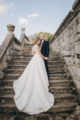 Gorgeous wedding couple walking on stone stairs near old castle in park. Stylish beautiful bride in...
