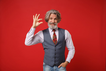 Confused elderly gray-haired mustache bearded man in classic shirt vest tie isolated on red wall...