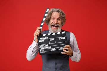 Laughing elderly gray-haired mustache bearded man in classic shirt vest tie isolated on red background. People lifestyle concept. Mock up copy space. Holding classic black film making clapperboard.