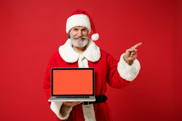 Elderly gray-haired mustache bearded Santa man in Christmas hat isolated on red background. New Year 2020 celebration concept. Mock up copy space. Hold laptop with blank screen, pointing finger aside.