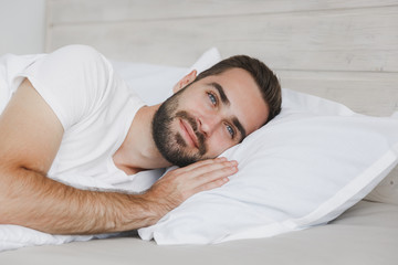 Fototapeta na wymiar Calm young handsome bearded man lying in bed with white sheet pillow blanket in bedroom at home. Smiling beauty male spending time in room. Rest relax good mood lifestyle concept. Mock up copy space.