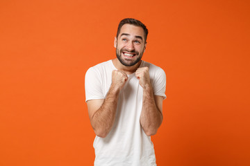 Excited overjoyed young man in casual white t-shirt posing isolated on bright orange wall background, studio portrait. People sincere emotions lifestyle concept. Mock up copy space. Clenching fists.
