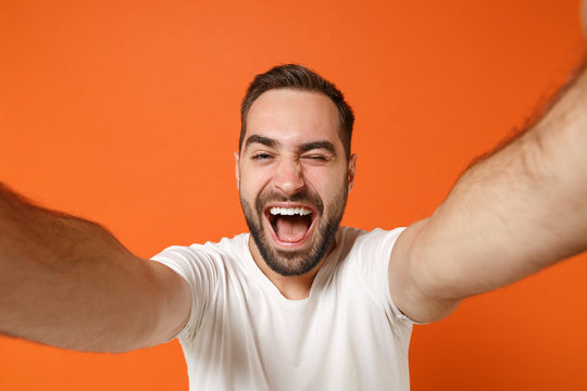 Close up of cheerful young man in casual white t-shirt posing isolated on orange background studio portrait. People lifestyle concept. Mock up copy space. Doing selfie shot on mobile phone, blinking.