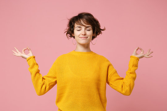 Young brunette woman girl in yellow sweater posing isolated on pastel pink background. People lifestyle concept. Mock up copy space. Hold hands in yoga gesture relaxing meditating keeping eyes closed.