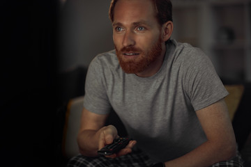 Portrait of bearded adult man watching TV at night while siting on couh in dark room and switching channels, copy space