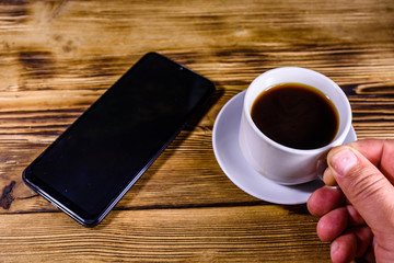 Fototapeta na wymiar Modern smartphone with blank screen on a wooden background and hand with cup of coffee