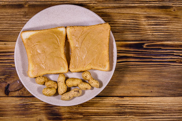 Glass jar with peanut butter and plate with sandwiches on a wooden table. Top view