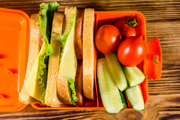 Lunch box with sandwiches, cucumbers and tomatoes on wooden table. Top view