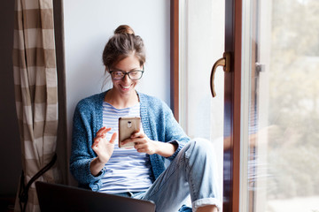 Holiday shopping online. Young woman makes purchases in the Internet. Freelancer pays by using smartphone on Black Friday. Happy girl is enjoying buying gifts and sitting by window with laptop at home - obrazy, fototapety, plakaty