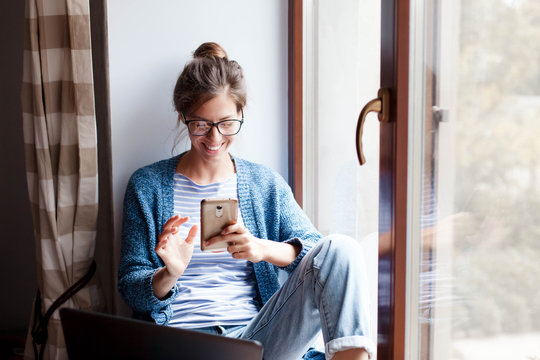 Holiday shopping online. Young woman makes purchases in the Internet. Freelancer pays by using smartphone on Black Friday. Happy girl is enjoying buying gifts and sitting by window with laptop at home