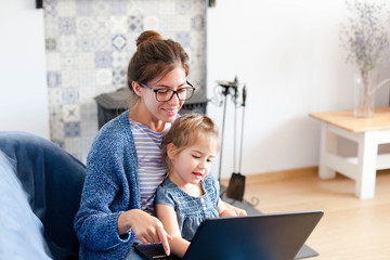 Holiday shopping online. Family make purchases in the Internet on Black Friday. Happy mother and child girl are enjoying buying gifts with laptop in cozy living room at home. Kid and woman are smiling