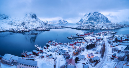 Aerial view of Reine in overcast day in winter in Lofoten islands, Norway. Moody landscape with blue sea, snowy mountains in clouds, rocks, village, buildings, red rorbu, cloudy sky. Top view. Travel