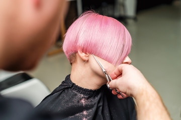 Professional hairdresser is cutting short pink hair of young woman in beauty salon.