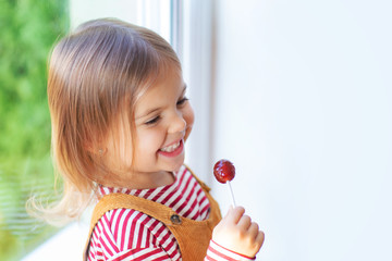 Portrait of cute toddler girl is sitting on windowstill with loly pop.