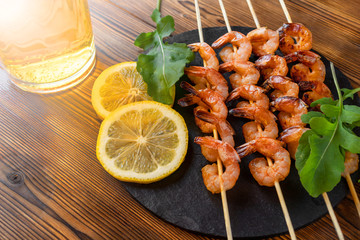 Shrimp fried on skewers with arugula, lemon on a black slate dish and beer in a glass on a wooden table