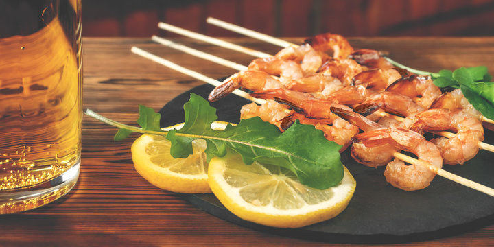 Shrimp fried on skewers with arugula, lemon on a black slate dish and beer in a glass on a wooden table