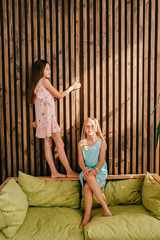Obraz premium Little blonde girl embracing legs of her girlfriend standing on bed with wooden wall on background.