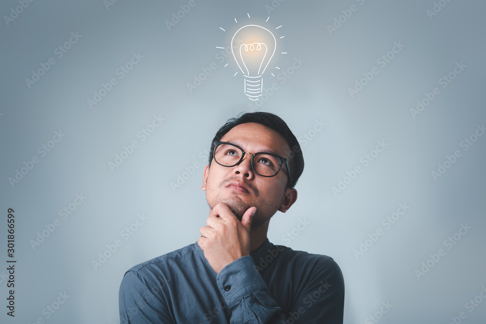 Aufkleber thinking asian man in glasses looking up with light idea bulb above head on gray background.creative