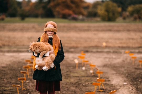 Redhead frekles blindfolded girl holding red cat in autumn field with lifeless landscape on background.