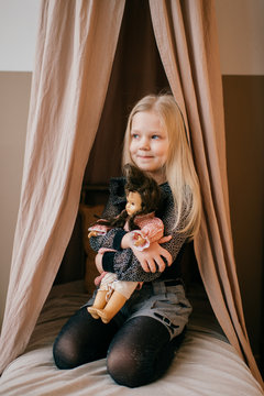 Beautiful little girl sitting on bed in wigwam and holding her loving doll in cozy room with decorations.