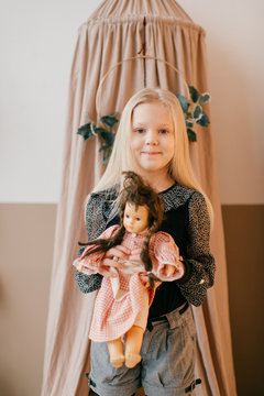 Beautiful little girl sitting on bed in wigwam and holding her loving doll in cozy room with decorations.