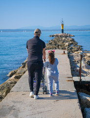 Dad with a small daughter and a baby in a stroller walks along the pier to the lighthouse in sunny...