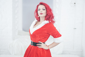pretty redhaired young stylish woman in velvet santa dress with fur posing on white room background near the cozy window