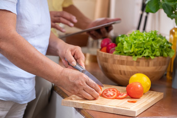 Asian senior wife slicing tomatoes on wooden cutting  board and using tablet computer to searching menu recipe.senior with technology lfiestyle.aging at home