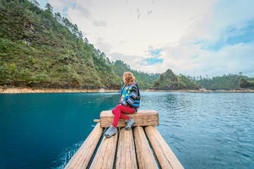 Young woman sitting on a wooden raft in the middle of a turqeuza blue lake admiring the landscape of wooded mountains on a cloudy day