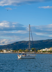 Sailing yacht, sailboat in the bay on city landscape and cloudy blue sky background. The boat is anchored in the bay with a calm sea. View from the sea to the coastal city. Roses, Catalonia, Spain.