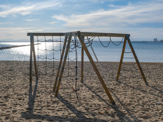 Childrens swing on the sandy beach on a background of blue sky and sea. Concept of family vacation. Summer day in Spain, Catalunya. Playground on beach.