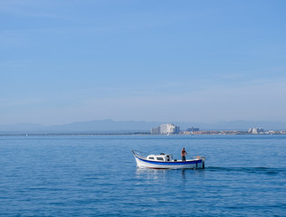 Fototapeta na wymiar Fisherman on a small boat at sea fishing on coastal city and blue sky background. Fishing in the bay by a city dweller. Calm water in sunny day. Roses, Catalonia, Spain. 