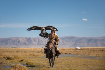 A proud young kazakh eagle hunter posing with his golden eagle on horseback on the backdrop of blue...