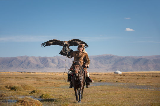 A proud young kazakh eagle hunter posing with his golden eagle on horseback on the backdrop of blue sky. Ulgii, Mongolia.