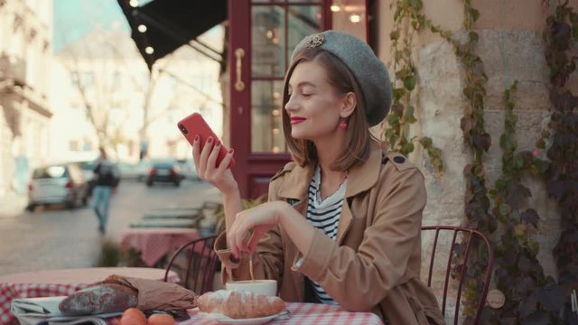 Happy young woman with red lipstick hat wearing stylish coat typing on phone during sunny day sitting in cafe on breakfast outdoors smile city fashion online slow motion