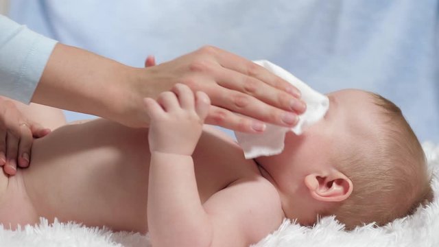 Mom wipes baby with wet wipe. Child lies on white blanket on blue background, mother wipes his face, neck, chest and abdomen with wet cloth. Slow motion