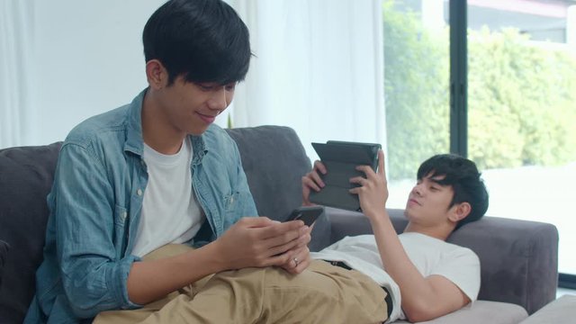 Young gay lgbtq couple using mobile phone and tablet at modern home. Asian lover male happy relax laugh and fun technology play games in internet together while lying sofa in living room concept.