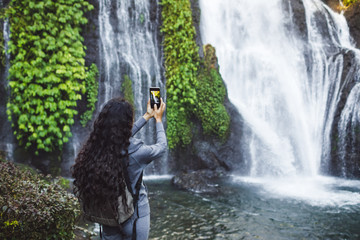 Fototapeta na wymiar Woman taking picture of waterfall Banyumala in Bali. Long curly brunette hair, backpack. Travel concept.