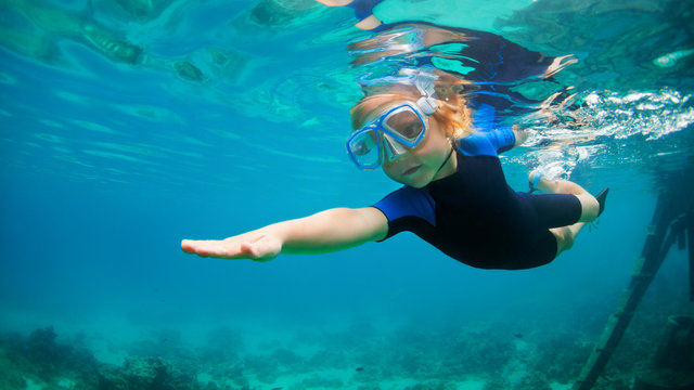Happy little kid in snorkeling mask and wetsuit jump and dive underwater in coral reef sea lagoon. Family travel lifestyle in summer adventure camp. Swimming activities on beach vacation with child.