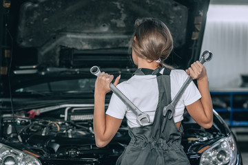 Young girl near the open hood of a black car in the garage. The girl is dressed in black jumpsuit and white t-shirt, she stands with her back to the camera. Girl holds up the wrenches