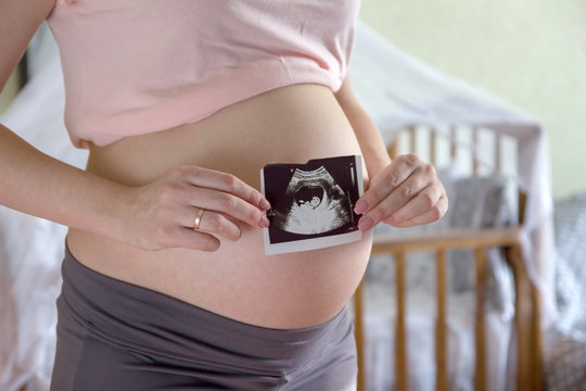 Big pregnant tummy and woman's hands holding ultrasound image of healthy unborn baby in the interior of the children's room. Maternity prenatal care and woman pregnancy concept.