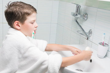 a boy in a white bathrobe in the bathroom brushes his teeth and washes his hands at the same time