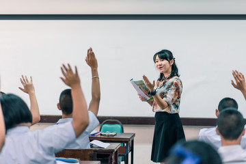 An smiling Asian female high school teacher teaches the white uniform students in the classroom by asking questions and then the students raise their hands for answers.