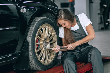 A beautiful girl in a white T-shirt and black jumpsuit is sitting near a black car removing the wheel in the service station