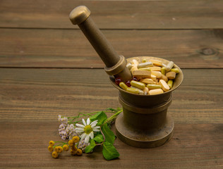 A copper mortar full of capsules and pills. Nearby lie medicinal flowers