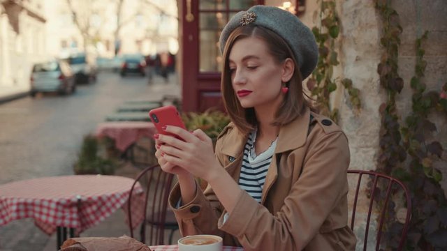 Charming woman with red lipstick hat wearing stylish coat typing on phone during sunny day sitting in cafe on breakfast outdoors city fashion online slow motion