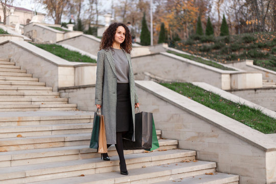 Full length picture. A cheerful young woman, with curly hair, elegantly dressed in her coat, goes down the stairs, holdings a lot of shopping bags, on a beautiful autumn day.